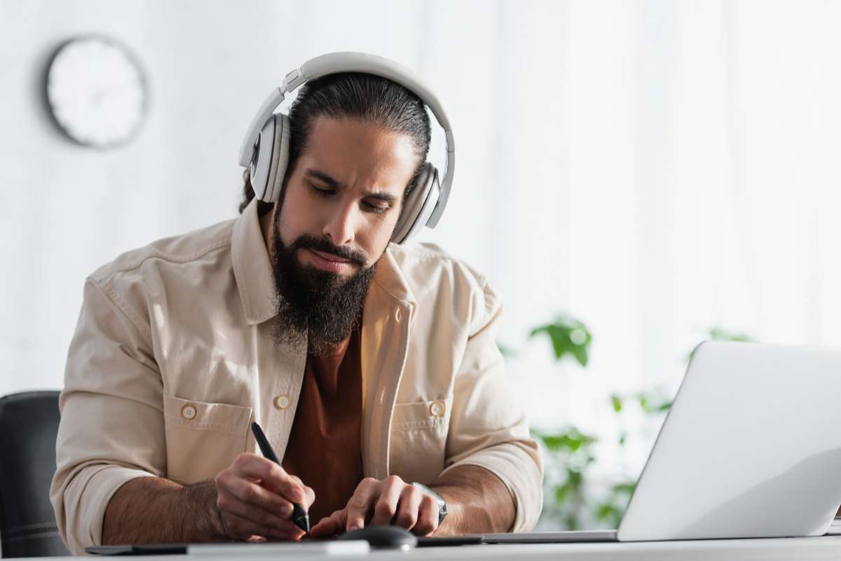 Hispanic designer in headphones drawing with stylus near blurred laptop (R) (S)